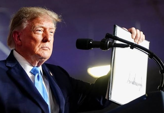 Man in a suit at a podium holding a large white document near a microphone