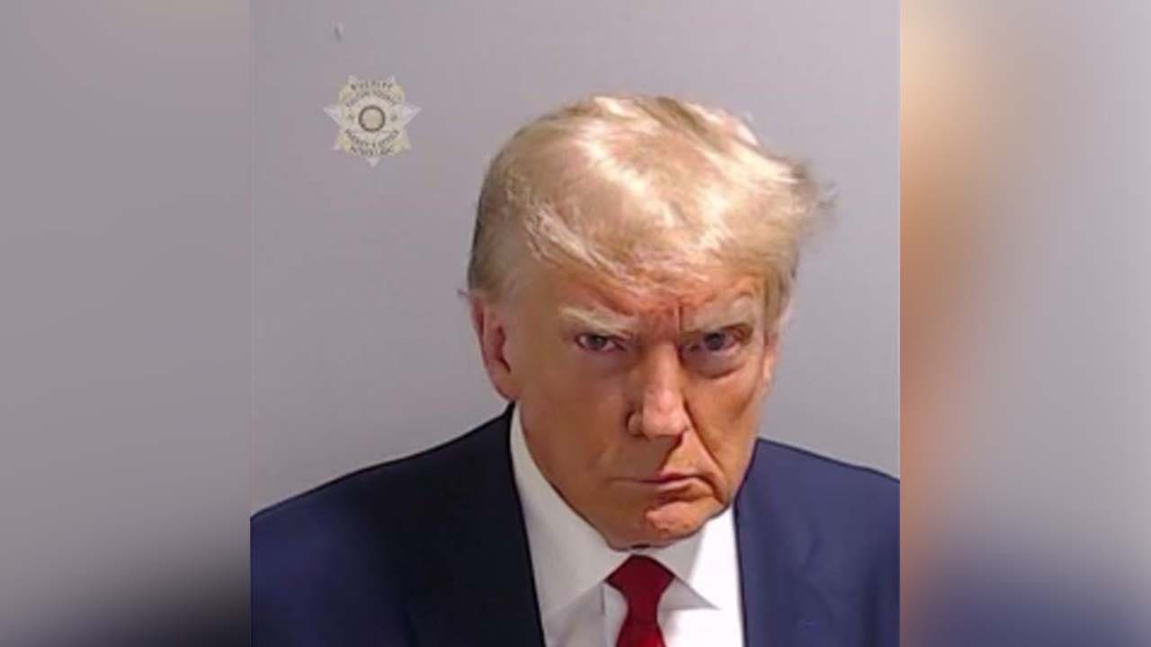 Mugshot-style photo of a stern-looking man in a dark suit and red tie against a plain gray wall