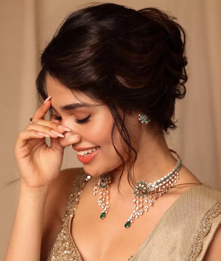 Krithi Shetty smiling gracefully in a champagne gold bridal saree and diamond-emerald necklace.