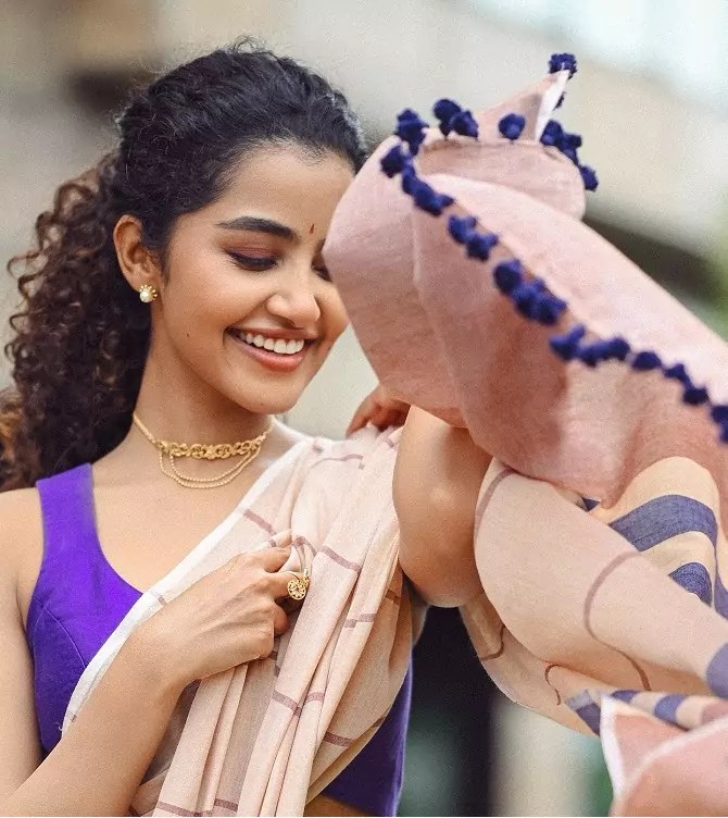 Anupama Parameswaran smiling in a beige saree with a sleeveless purple blouse.