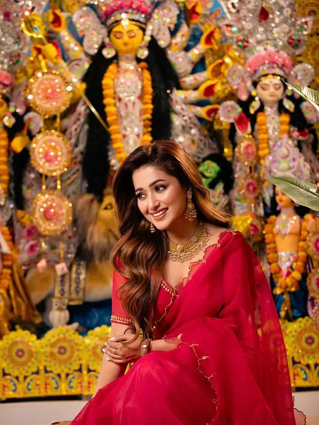 Sudipta Bakshi seated gracefully before Durga idol, radiating festive elegance in red attire.