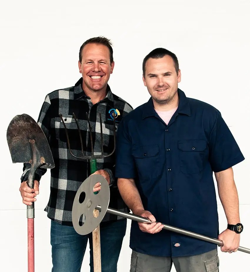 Two men stand side by side smiling, holding large tools. The man on the left wears a plaid shirt and holds a shovel and pitchfork. The man on the right wears a navy shirt and holds a metal tool with a circular end.