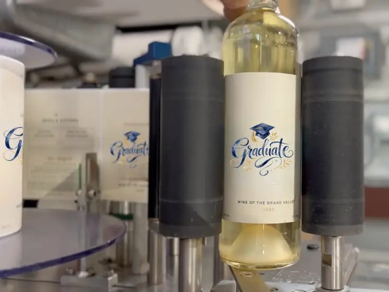 A bottle of white wine with a Graduate label and graduation cap icon moving through an industrial labeling machine.