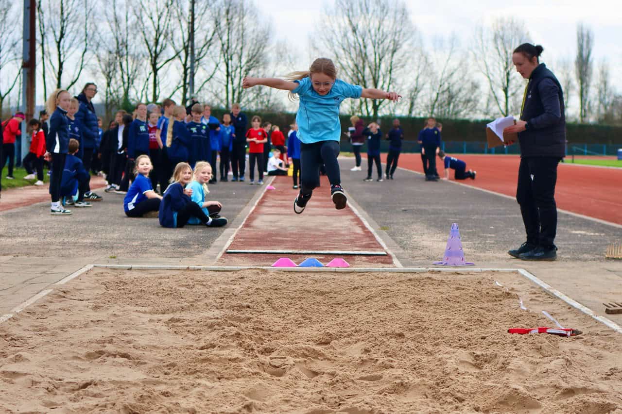 Young girl in blue jumpsuit competing in long jump at LKS2 Athletics Tournament.