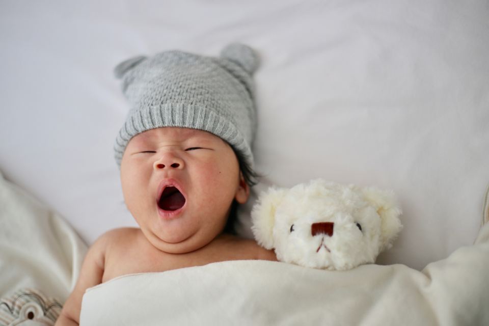 Baby yawning in their crib. A stuffed bear is tucked in next to the baby