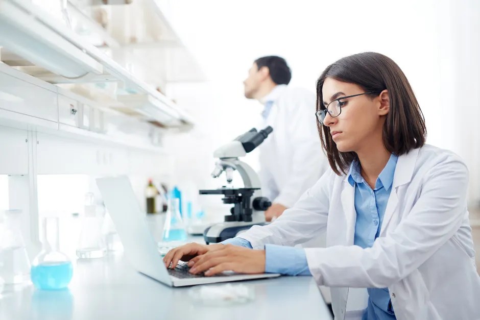 Woman scientist writing on her laptop in the laboratory with male colleague in the background