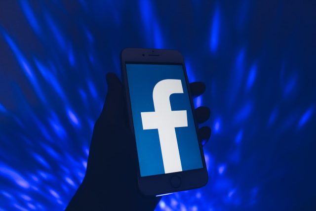 Illustration of a silhouette of a hand holding a phone with the Facebook logo on it.