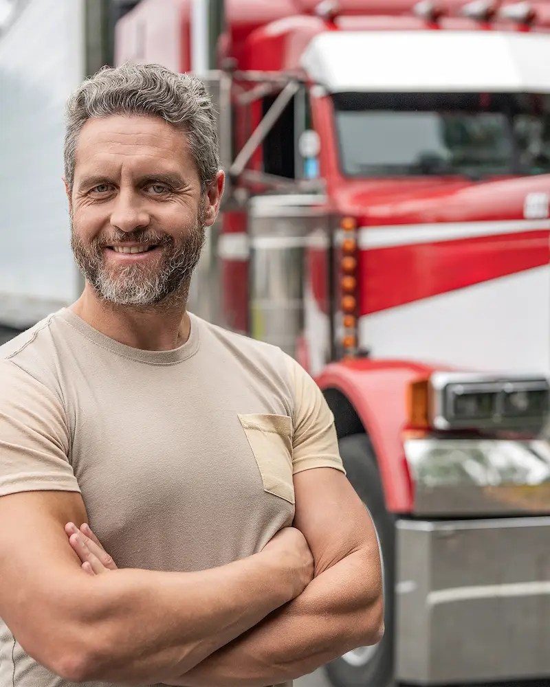 CDL truck driving school instructor standing with his arms crossed in front of a red 18 wheeler.