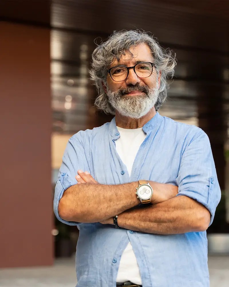 Older college professor with his arms crossed standing in front of a university building.