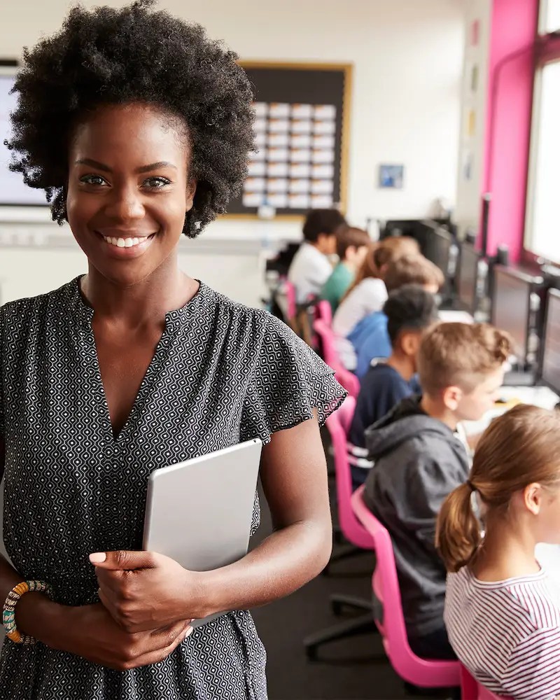 Smiling computer science teacher holding a tablet in a classroom filled with children working on desktops computers.