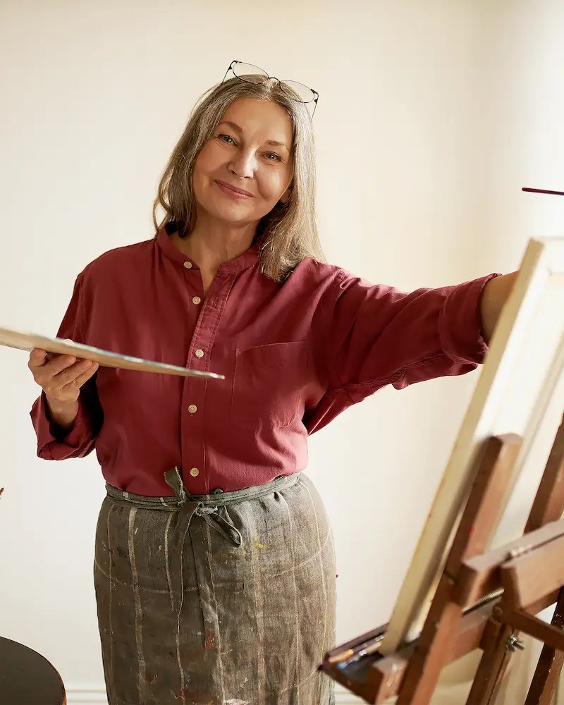 Mature woman painting on a canvas at an easel, holding a brush and wearing a paint-stained apron in an art school studio.