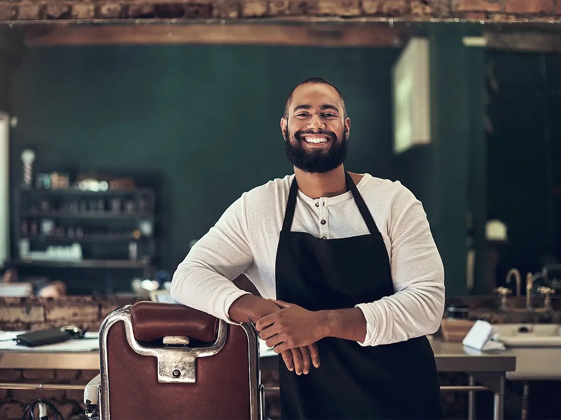 Smiling, male barber in a black apron leaning his arm on a barber chair in a hair salon.