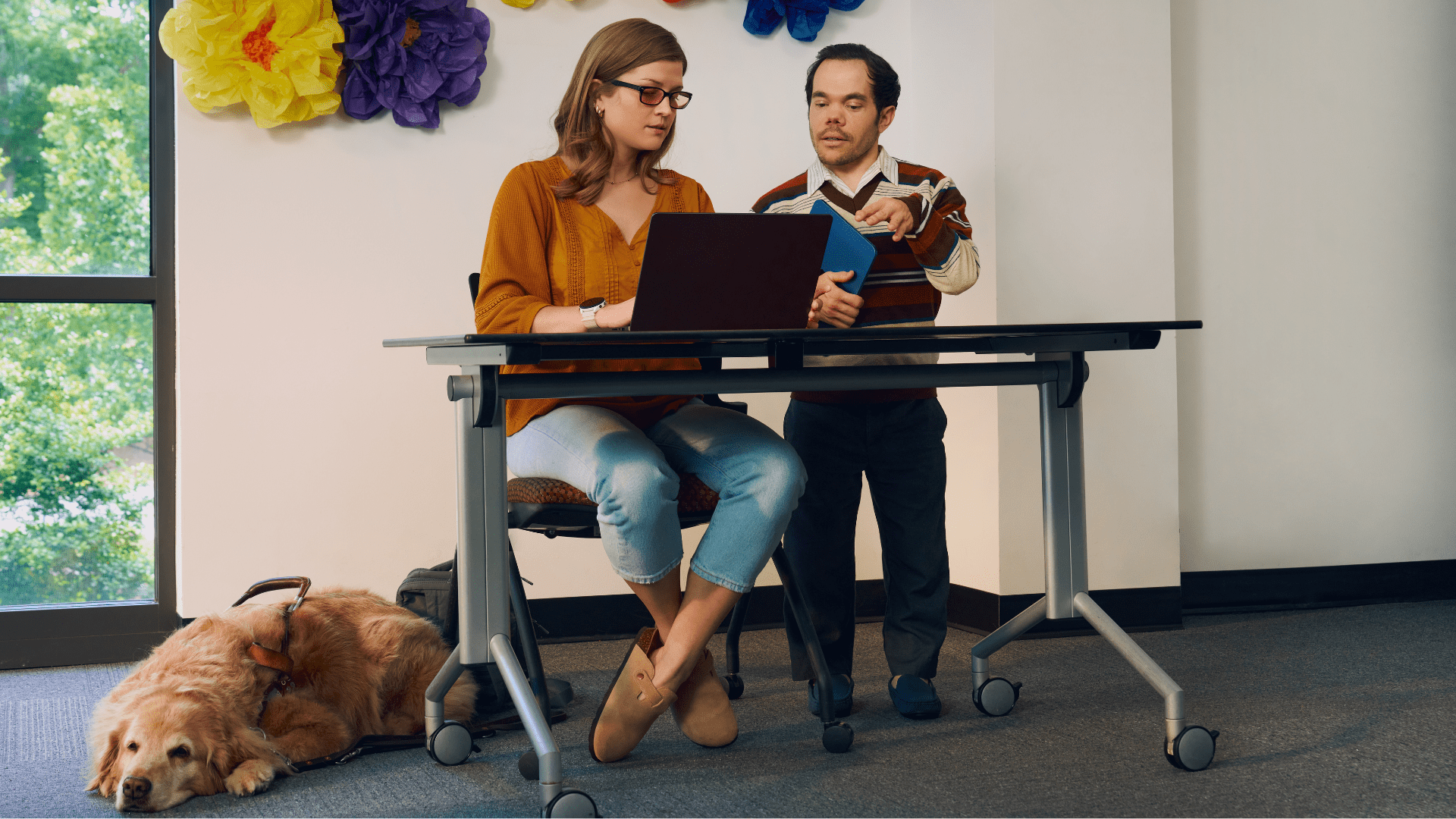 A blind, white woman wearing glasses with red lenses sitting at a lecture table, chats with her professor, a white man with medium brown hair and light brown eyes who has dwarfism, who is standing next to her. They are both looking intently at the student's laptop. A golden retriever service dog wearing a guide harness lays on the floor next to her. The student has dark blonde hair and is wearing an orange blouse and jean capris. A large flower sculpture hangs on the wall behind the lecture table. Next to the sculpture is a large window showing many large trees.