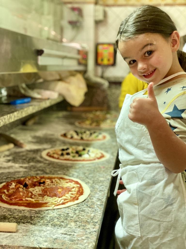 A young girl gives a thumbs up while making fresh pizza, a classic culinary experience during a family trip to Italy.