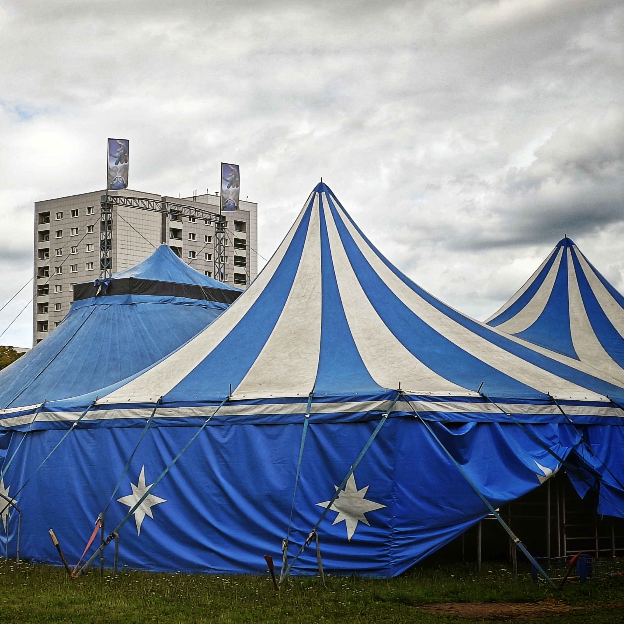 Blau-weiße Zirkuszelte vor grauen Hochhäusern und grauem Himmel.