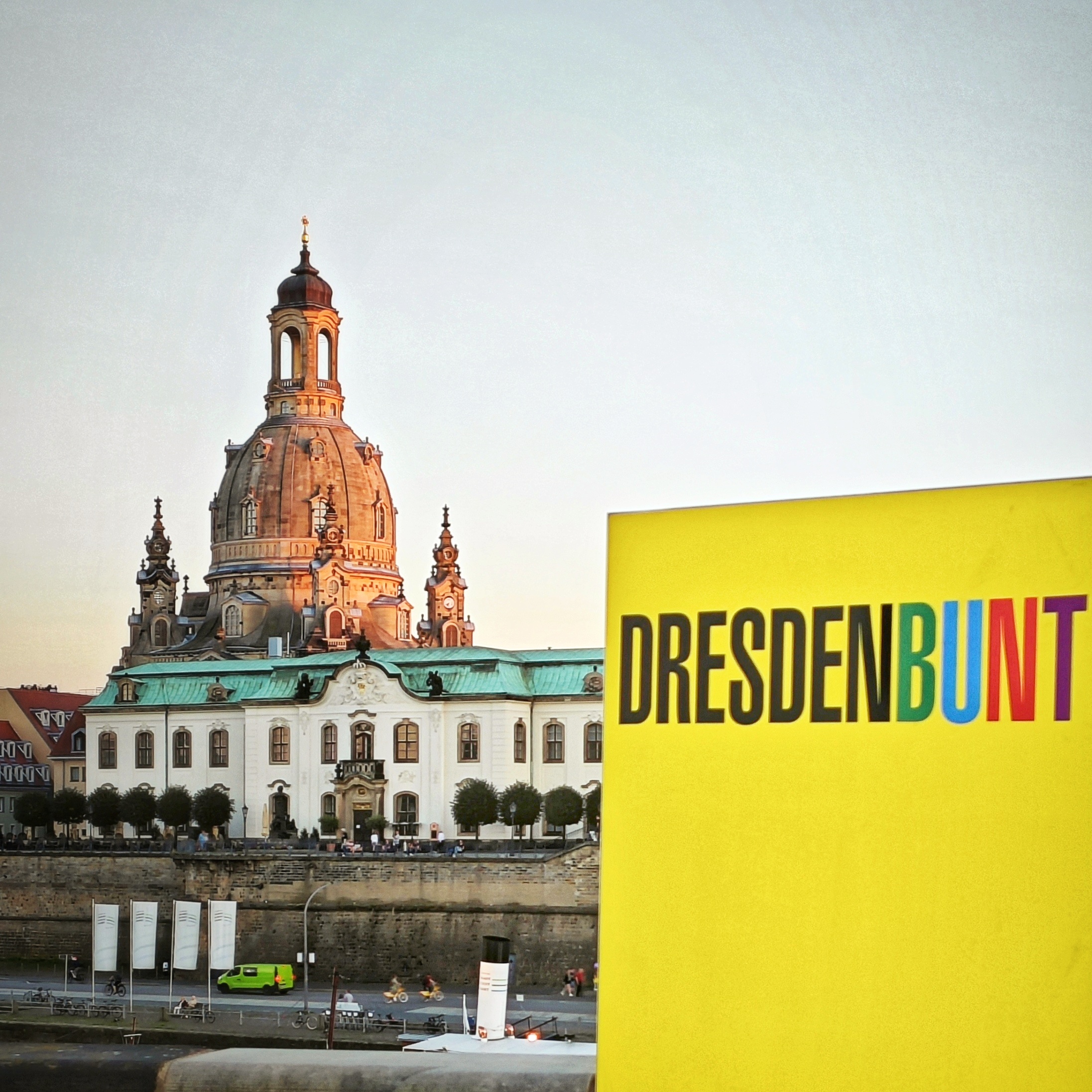 Die Frauenkirche von Dresden im Hintergrund. Im Vordergrund ein gelbes Schild, auf dem DRESDENBUNT steht. Die Buchstaben von BUNT sind grün, blau, rot und violett gedruckt.