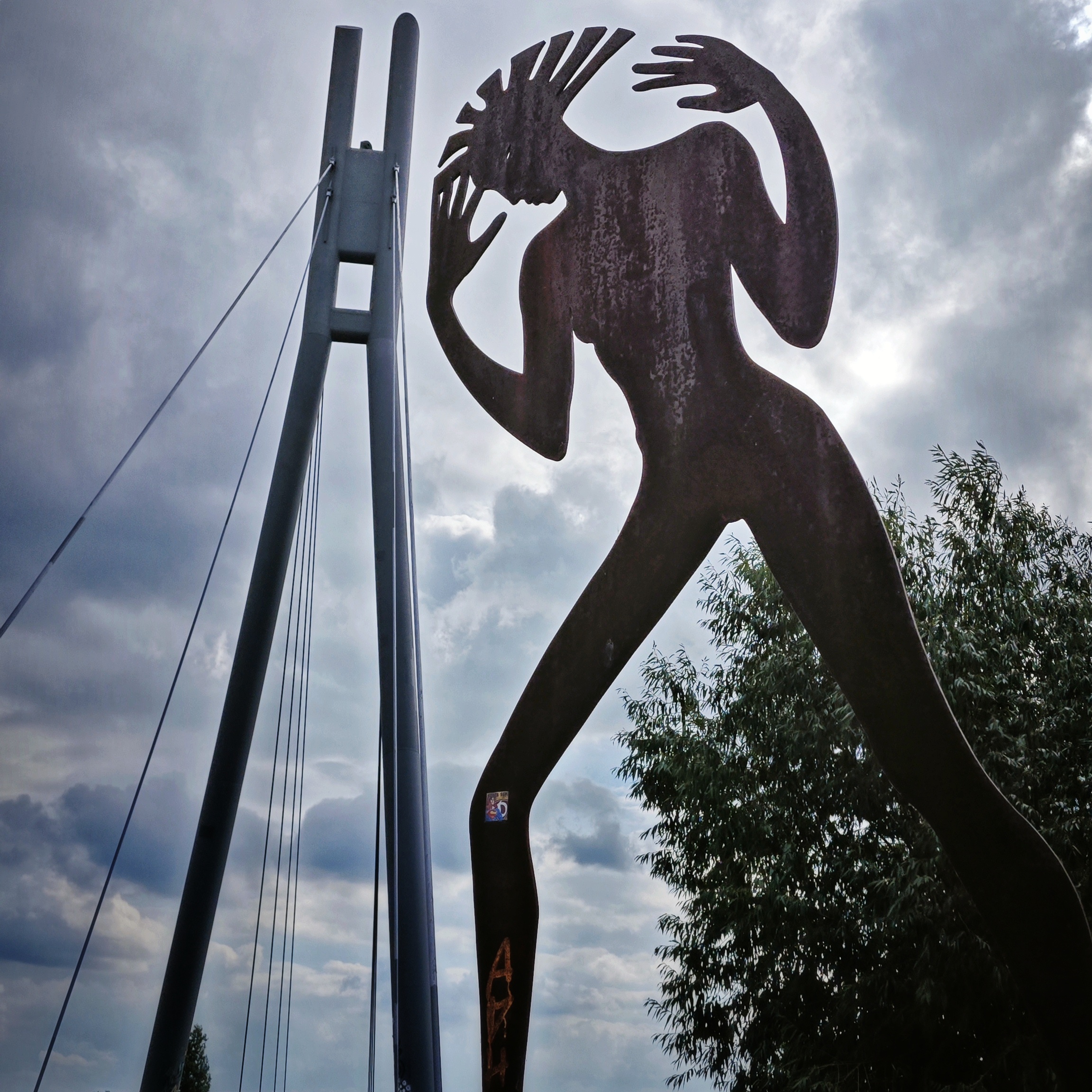 Stahlskulptur „Undine kommt“ von Angela Hampel an der Molenbrücke in Dresden-Pieschen nach der Erzählung „Undine geht“ von Ingeborg Bachmann. Die dünne Frauengestalt mit sonnenstrahlartigen Haaren hebt die Hände zum Kopf und scheint - perspektivenbedingt - fast so hoch wie die Molenbrücke zu sein.