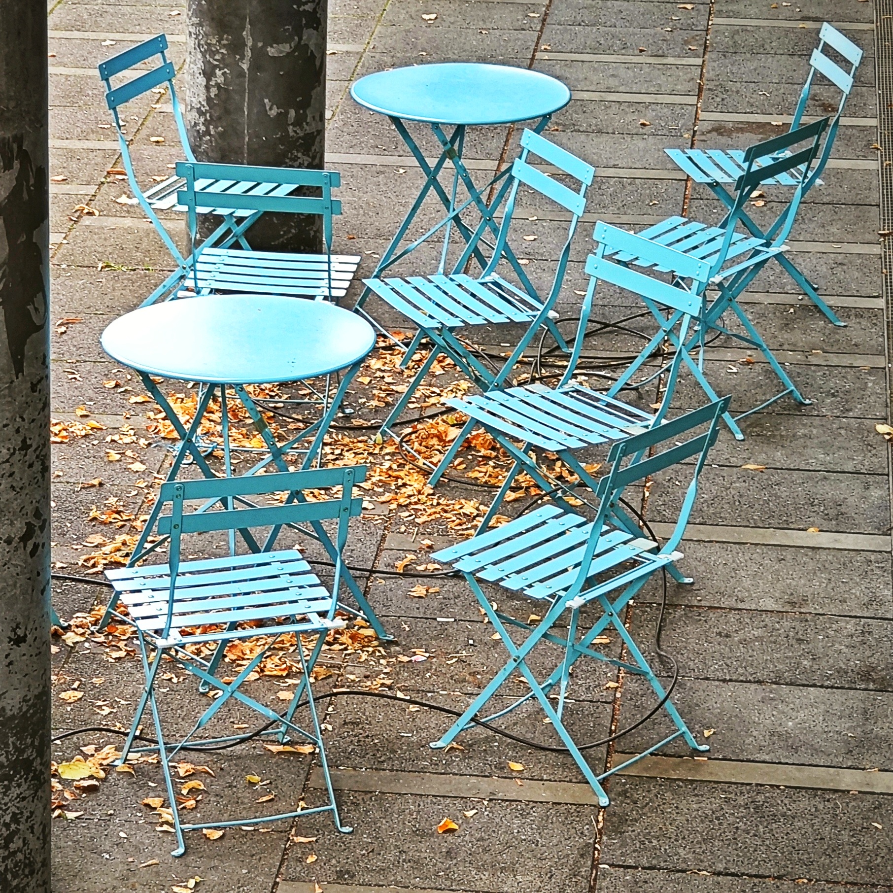 Eine hellblaue Sitzgruppe aus zwei runden Tischen und acht Klappstühlen steht wild angeordnet auf Betonplattenboden. Unter den Tischen und Stühlen liegt etwas gelbes Herbstlaub.