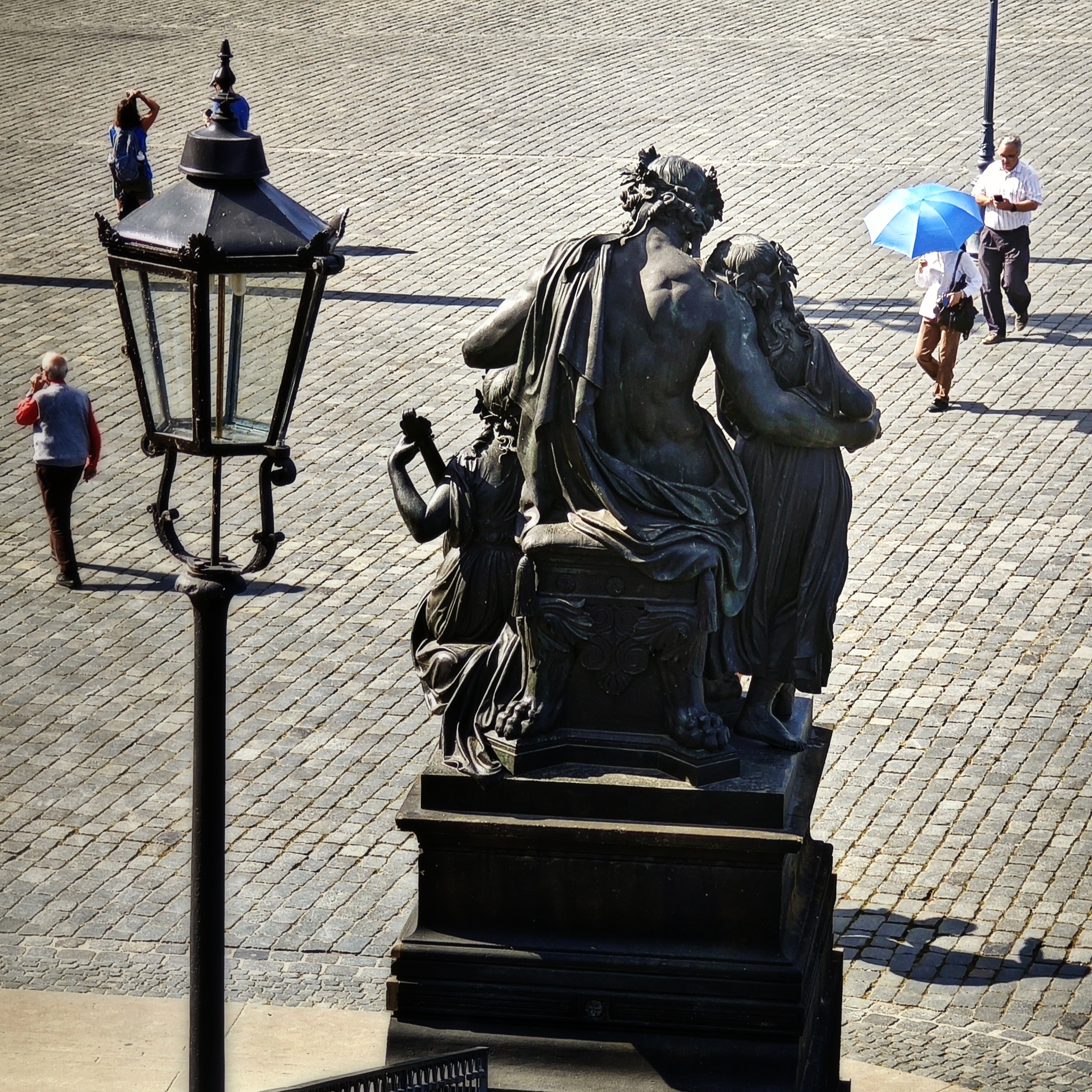 Bronzestatue "Abend" der Figurengruppe "Vier Jahreszeiten" des Bildhauers Johannes Schilling am Fuß der Freitreppe zur Brühlschen Terrasse von hinten betrachtet. Eine Laterne links. Vier Personen auf dem Schlossplatz verteilt, darunter eine mit aufgespanntem, blauem Sonnenschirm.