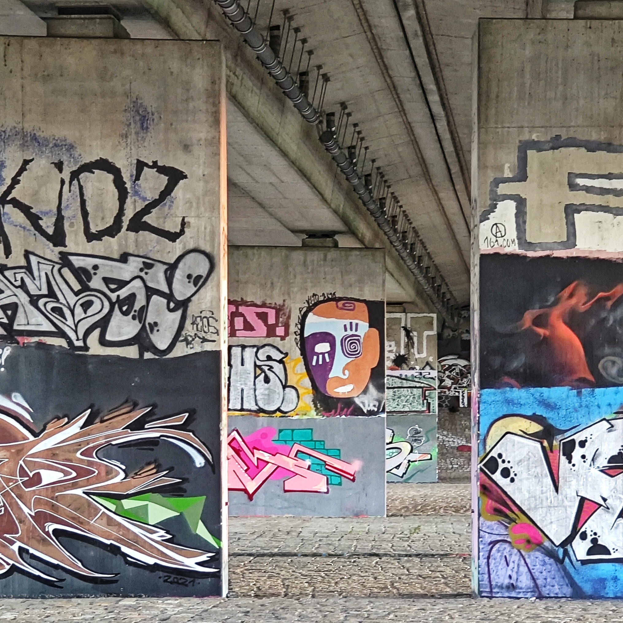 Graffiti: Teilansicht der Brückengalerie mit dem bunten Avangardekopf im Hintergrund