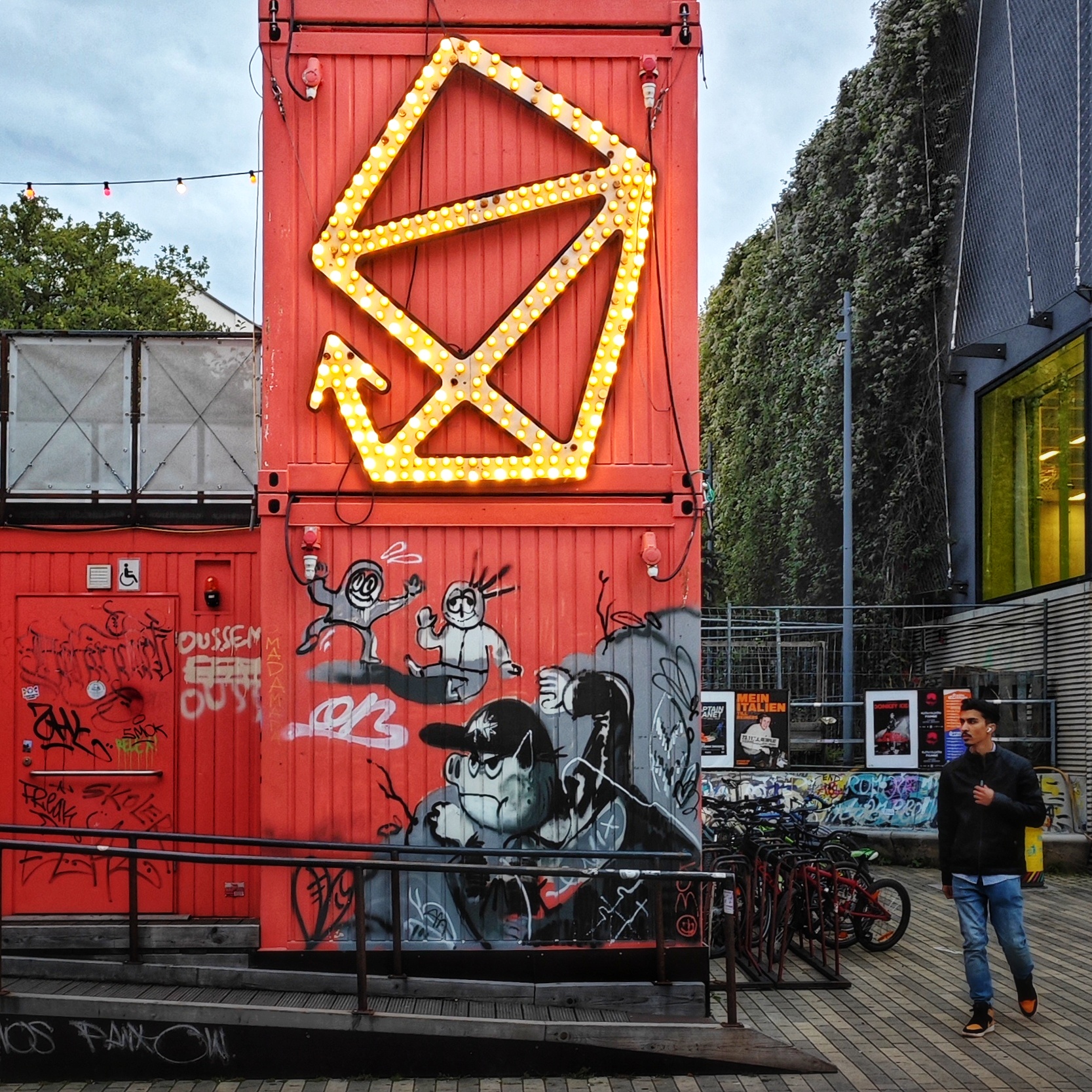 Das Logo des Dresdner Kulturvereins scheune e.V. - fast das Haus vom Nikolaus, nur die linke Seite ist statt geschlossen ein Pfeil nach oben - auf rotem Blechcontainer. Den unteren roten Blechcontainer zieren grau-schwarze Graffiti.