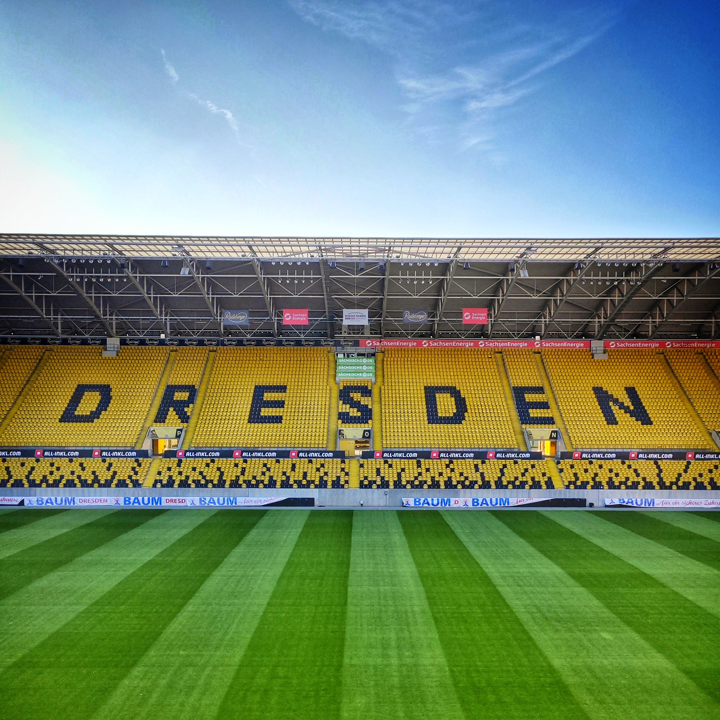 Innenansicht aus dem Rudolf-Harbig-Stadion auf die Gegengerade. Der schwarze Schriftzug DRESDEN in großen Buchstaben hebt sich von den sonst gelben Sitzen ab.