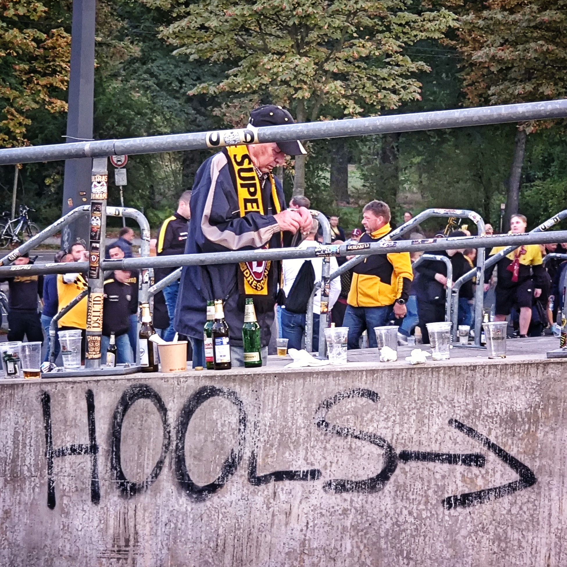 Am Dresdner Rudolf-Harbig-Stadion. Einige Dynamo-Fans mit gelb-schwarzer Fankleidung. Ein älterer Herr mit gelb-schwarzem Dynamo-Schal geht nach rechts die Treppe hinauf. Im Vordergrund weist ein Graffito darauf hin, dass es nach rechts zu den HOOLS geht.