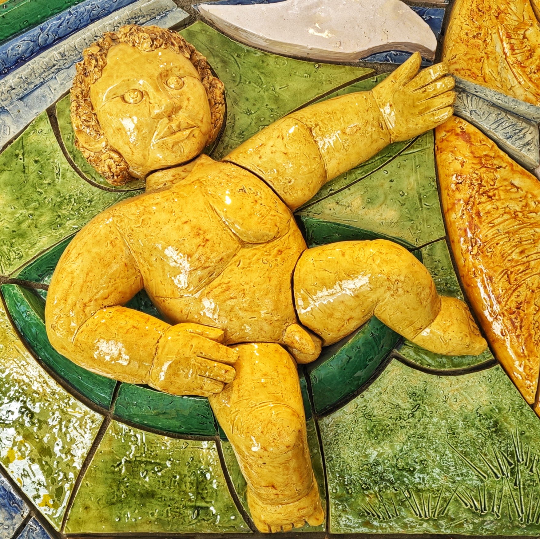 Teil des Keramikreliefs des Keramikers Dieter Graupner (1983), das bis zur Schließung der Rosen-Apotheke im dortigen Innenraum angebracht war. Der Teil zeigt einen gelben nackten Knaben auf grünem Grund.