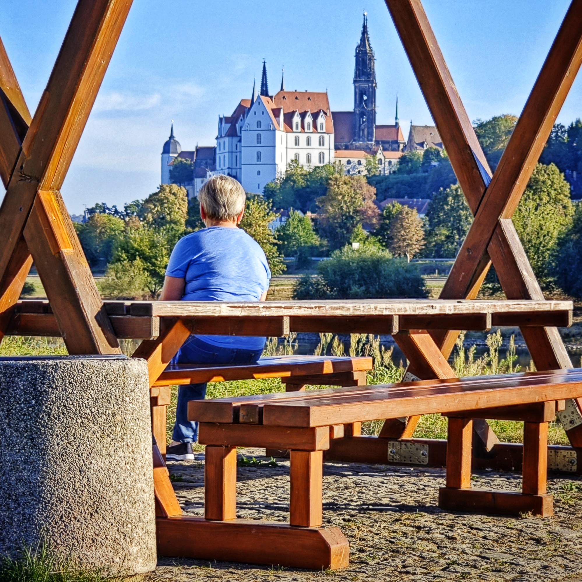 Eine Person mit weißen Haaren sitzt auf einer Holzbank mit Holzdach und schaut auf den Meißner Dom am jenseitigen Elbufer.