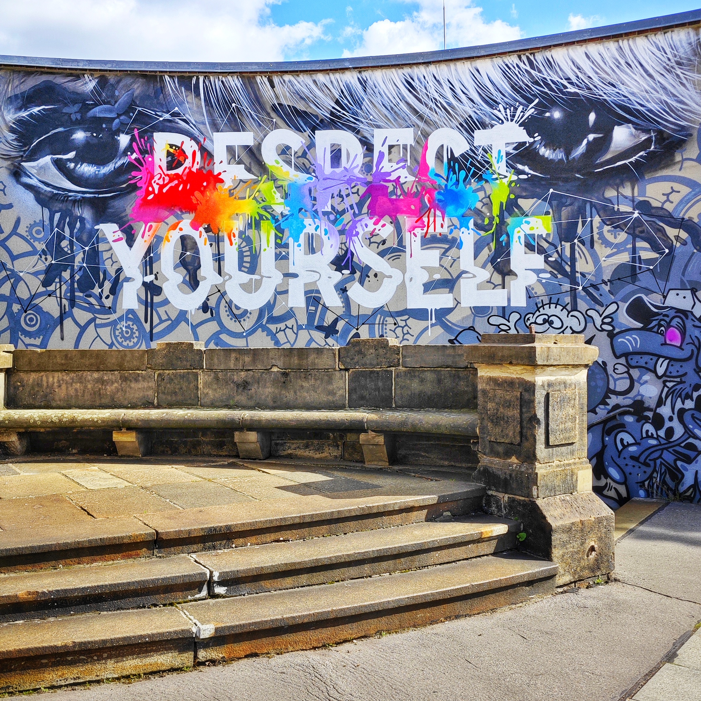 Graffiti an einer Wand vor einer kleinen Steintreppe. Das Graffiti zeigt zwei große Augen, vor denen in Großbuchstaben RESPECT YOURSELF steht. Der Slogan ist mit bunten Farbspritzern garniert.