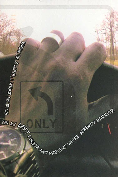 A close-up of a hand on a steering wheel, wearing a ring, with a blurred background of trees.