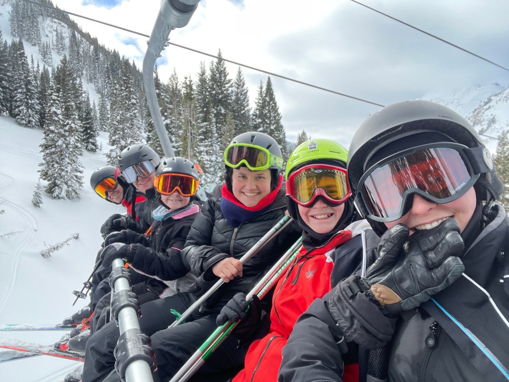 A group of six people, all wearing ski helmets and goggles, smiling while riding a ski lift in a snowy landscape with pine trees and mountains in the background.