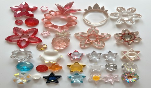 Coleção diversificada de formas pré-cortadas de acrílico para bijuterias dispostas sobre fundo branco, incluindo flores vazadas em tons de vermelho, rosa e transparente, estrelas coloridas e transparentes em diversos tamanhos, bolinhas com miolo colorido, passantes redondos, e peças decorativas em cores vibrantes como azul turquesa, amarelo, vermelho e rosa, ideais para confecção de colares e pulseiras artesanais.