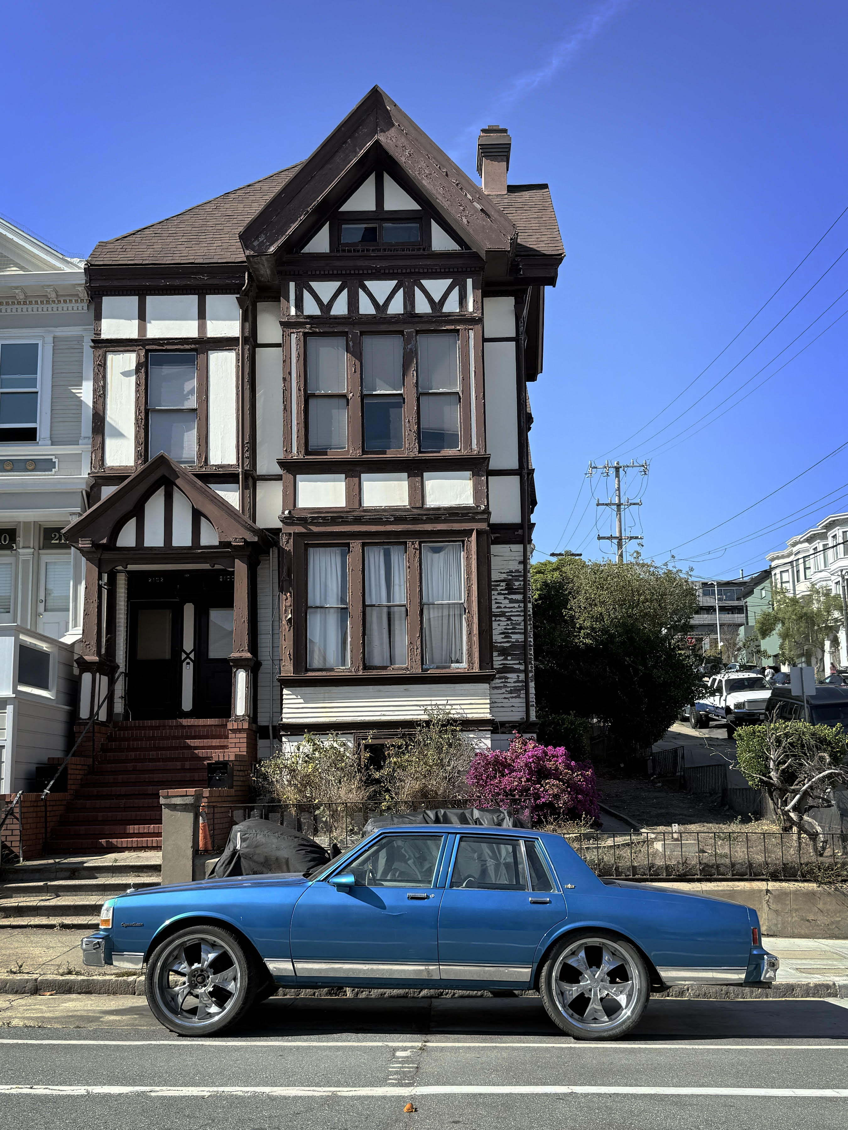 An electric blue sedan parked in front of a stately Tudor style house on a cloudless San Francisco day