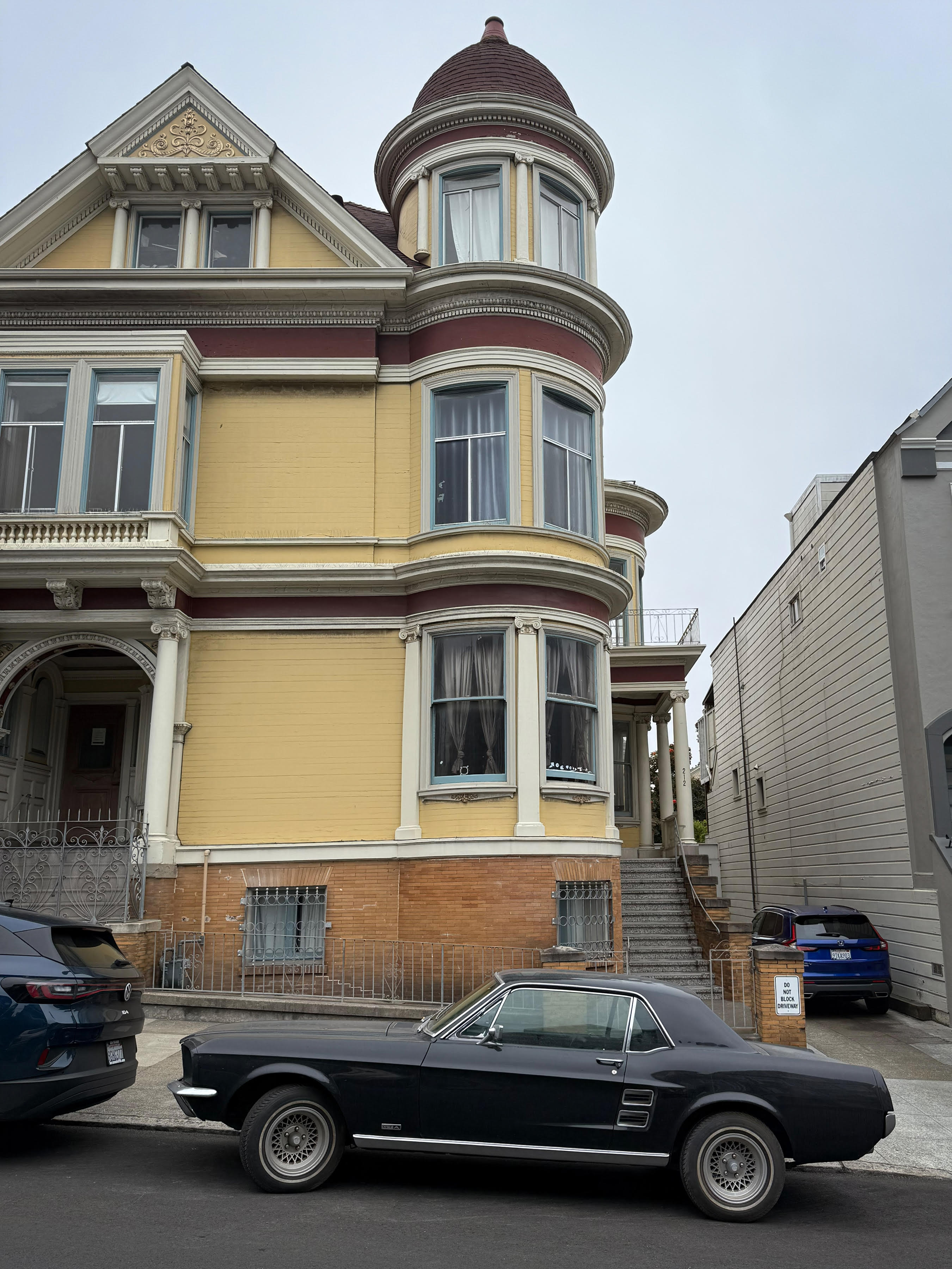 A black Mustang parked in front of a golden yellow Victorian house with a turret on a San Francisco city street