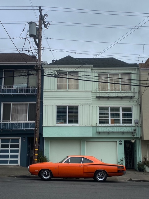 A flame-orange muscle car parked in front of a mint green building in San Francisco on a cloudy day