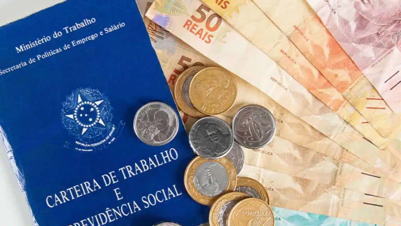 Imagem de uma carteira de trabalho da CLT, moedas e cédulas de dinheiro.