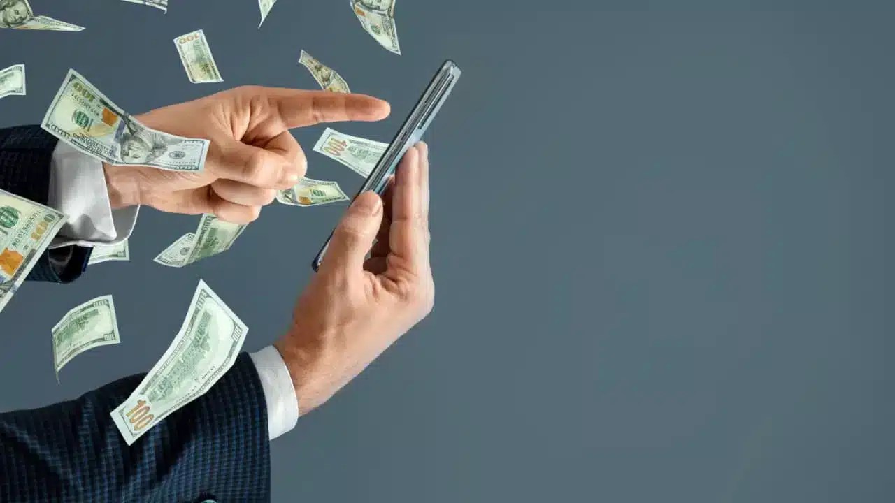 Celular com notas de dinheiro saindo da tela representando contas renumeradas de bancos.
