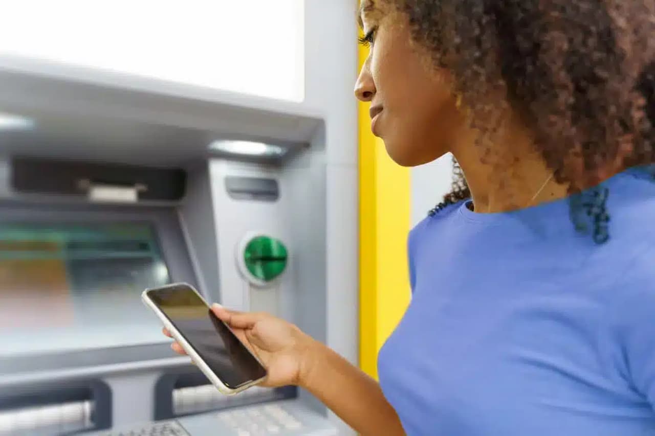 Imagem de mulher mexendo em aplicativo de banco, em frente a caixa eletrônico. DOC e TEC