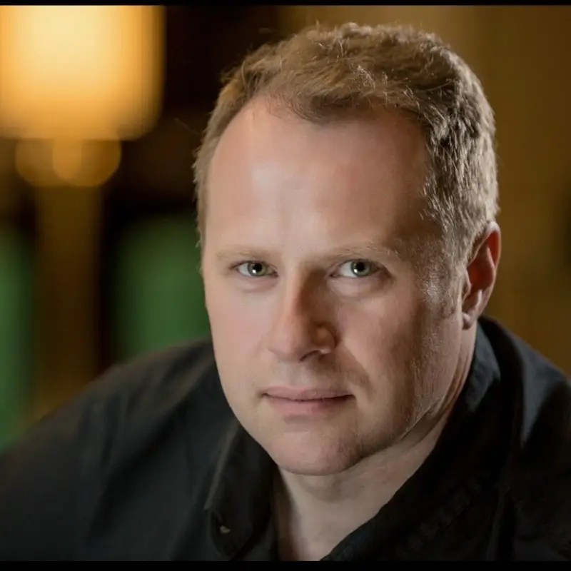 A man with short light brown hair wearing a black shirt, photographed in a warmly lit setting.