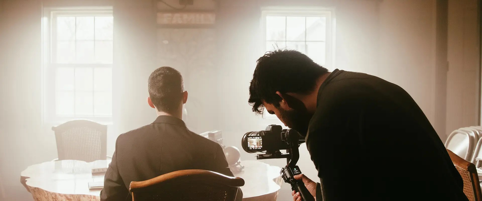 A videographer films a seated person from behind in a warmly lit room with two windows.