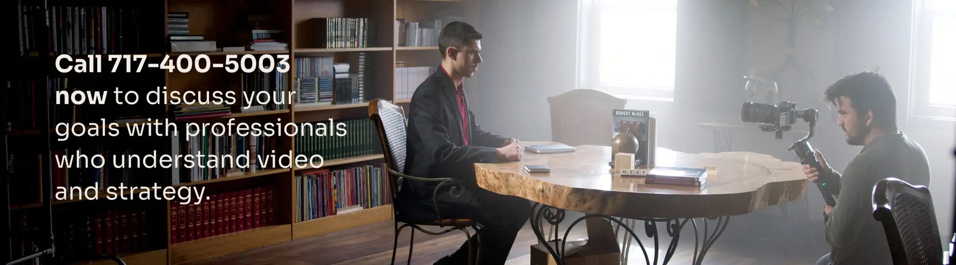 A man in a suit sits at a wooden table while a videographer films him in a library-style room.