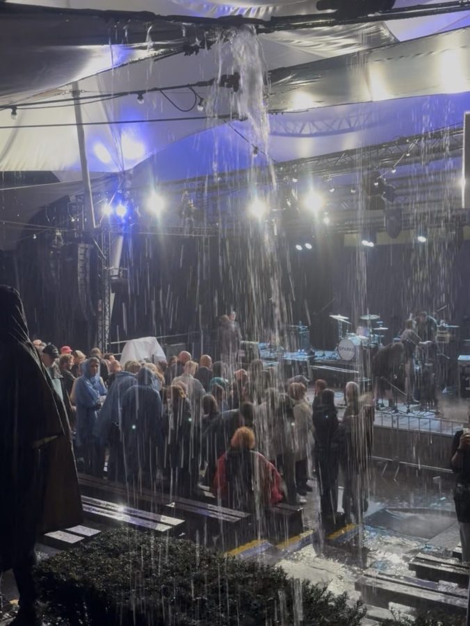 Rain pours in sheets off the edge of a white canopy at an outdoor concert, forming a curtain of water in front of the audience. Beyond it, people in ponchos stand watching an empty, brightly lit stage with a drum kit bearing the Sparks logo.