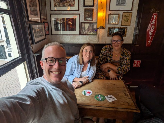 Three people pose for a selfie at a small wooden table in a traditional brown café, beer mats scattered in front of them. The walls behind are covered in framed prints and photographs, with a window onto a cobbled street to the left.