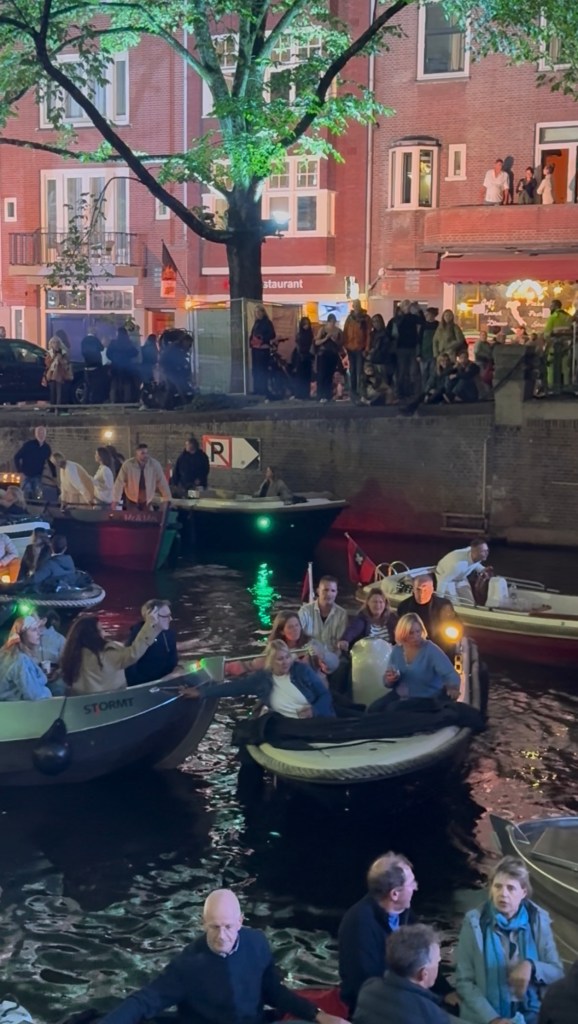 Open boats crowd a canal at night, packed with people watching a concert, with a lit lamp and Amsterdam flags among them. Crowds line the quayside above beside a row of red-brick buildings and a lit restaurant.