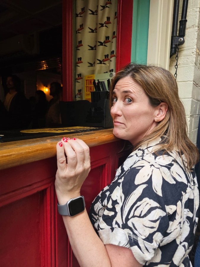 Sharon leaning on the counter of an outdoor bar hatch, pulling a rueful face at the camera, red polka-dot nails on the wood, tropical-bird curtain in the serving window behind.