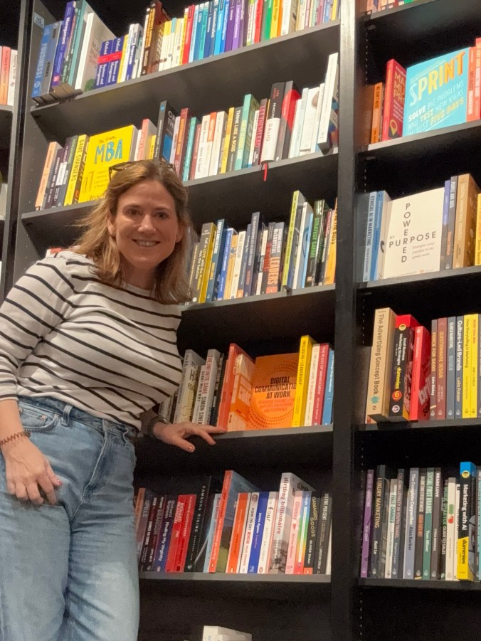 Sharon standing beside a business bookshop shelf, hand resting under a copy of Digital Communications at Work, shelved face-out among other management titles.