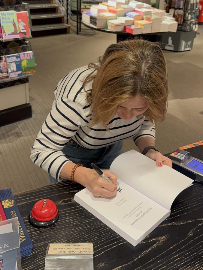 Sharon bent over a bookshop counter signing the title page of Digital Communications at Work, a service bell and stacks of new releases beside her.