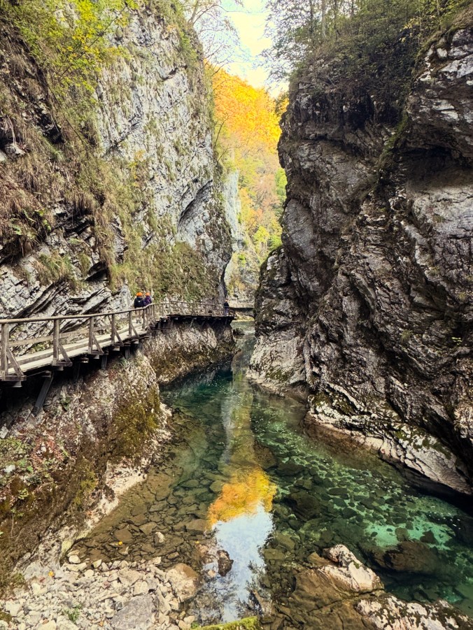 A wooden boardwalk clings to a sheer grey cliff in a narrow gorge. Two walkers in orange helmets head along it. Below, a still, clear green pool reflects a blaze of yellow autumn trees.