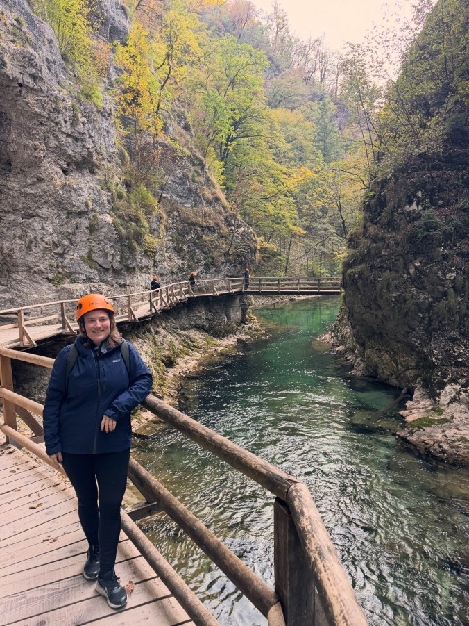 Sharon, in an orange safety helmet and navy jacket, smiles and leans on a wooden railing above a turquoise river. The boardwalk winds on behind her between steep, tree-covered rock walls.